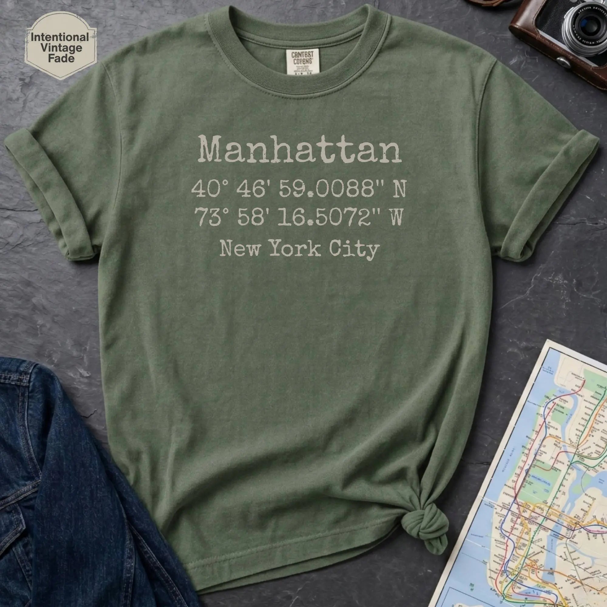 Moss green Comfort Colors t-shirt featuring Manhattan, NY coordinates and New York City text in a minimalist typewriter-style design.