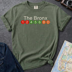Moss green Comfort Colors t-shirt featuring a minimalist Bronx subway line graphic with The Bronx text and colorful NYC train icons.