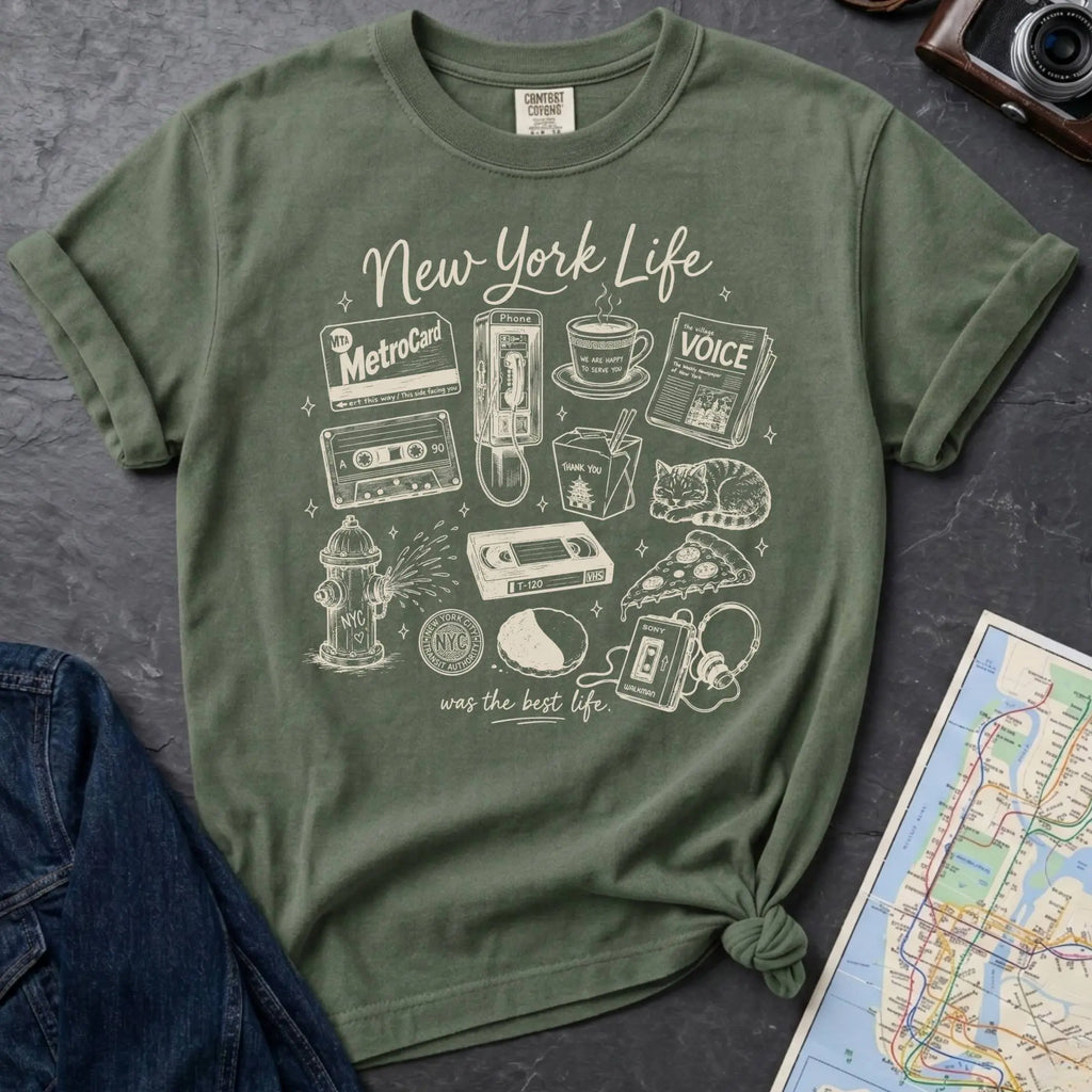 Moss green Comfort Colors t-shirt featuring a New York Life Was the Best Life graphic with hand-drawn NYC nostalgia icons including a MetroCard, cassette tape, pay phone, coffee cup, newspaper, pizza slice, fire hydrant, Walkman, and bodega cat.