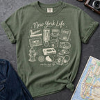 Moss green Comfort Colors t-shirt featuring a New York Life Was the Best Life graphic with hand-drawn NYC nostalgia icons including a MetroCard, cassette tape, pay phone, coffee cup, newspaper, pizza slice, fire hydrant, Walkman, and bodega cat.