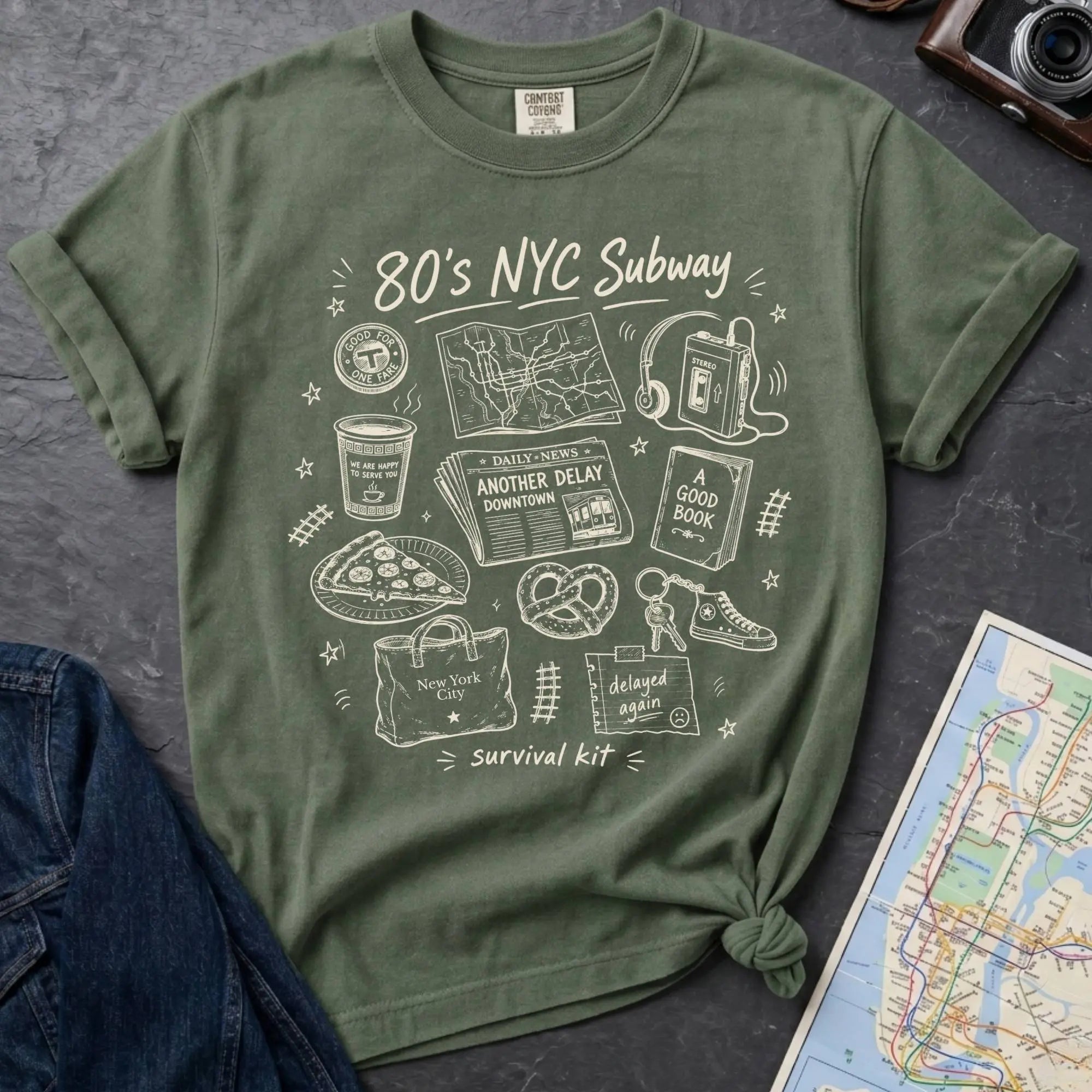 Moss green t-shirt with '80s NYC Subway Survival Kit' graphic design on a dark surface with a map and denim jacket.