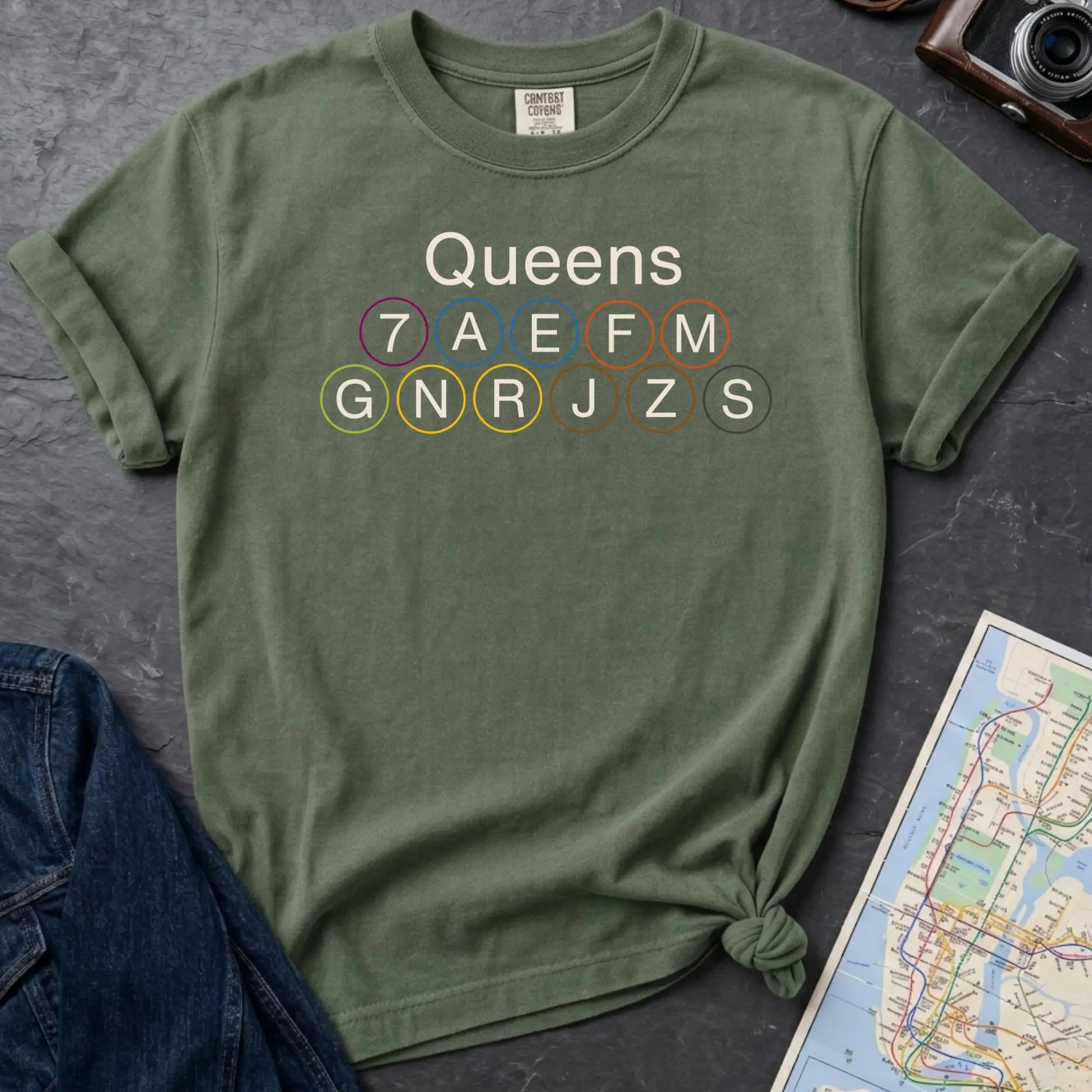 Moss green Comfort Colors t-shirt featuring a minimalist Queens subway line graphic with Queens text and classic NYC train icons.