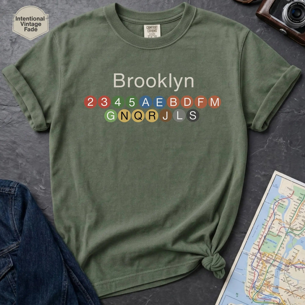 Moss green t-shirt with 'Brooklyn' and subway line numbers on a dark surface with a map and camera.