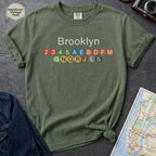 Moss green t-shirt with 'Brooklyn' and subway line numbers on a dark surface with a map and camera.