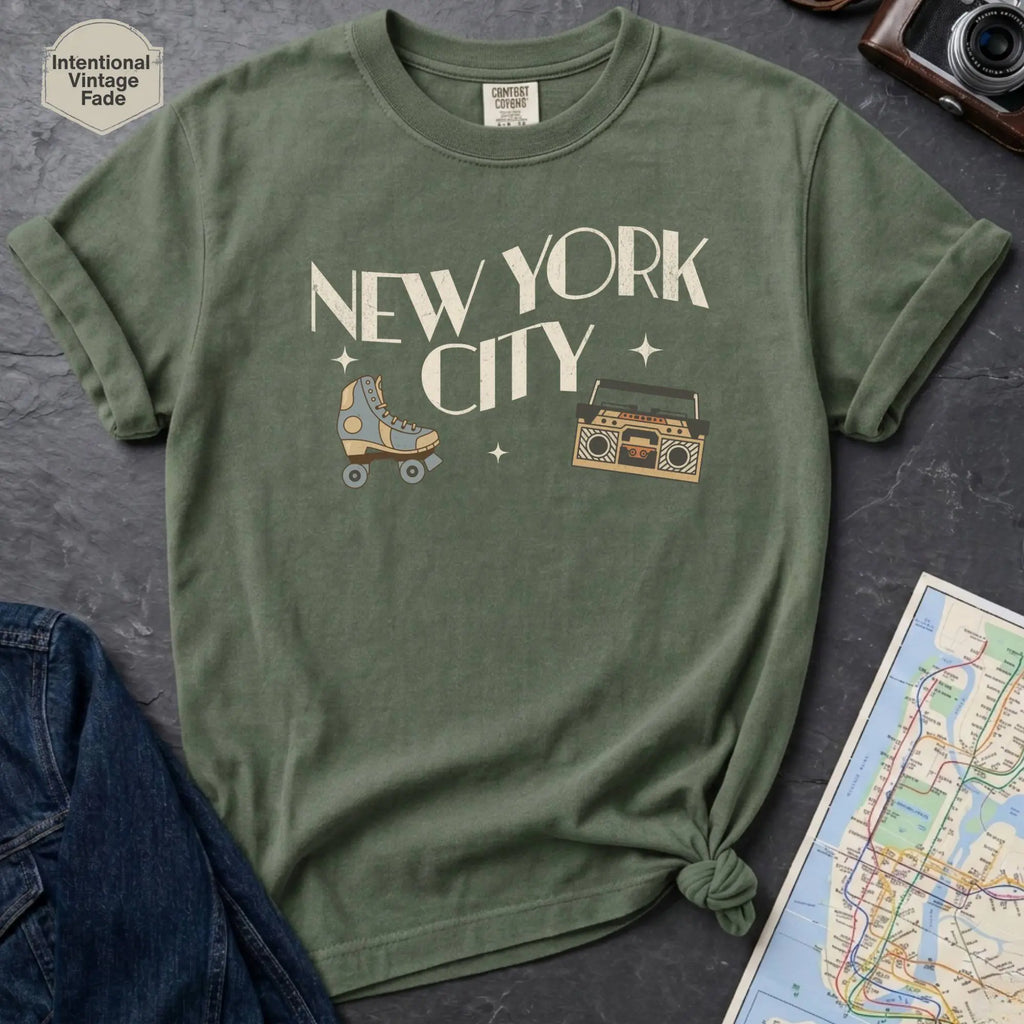 Moss green t-shirt with 'New York City' text and a roller skate and boom box on a dark surface with a map and camera.