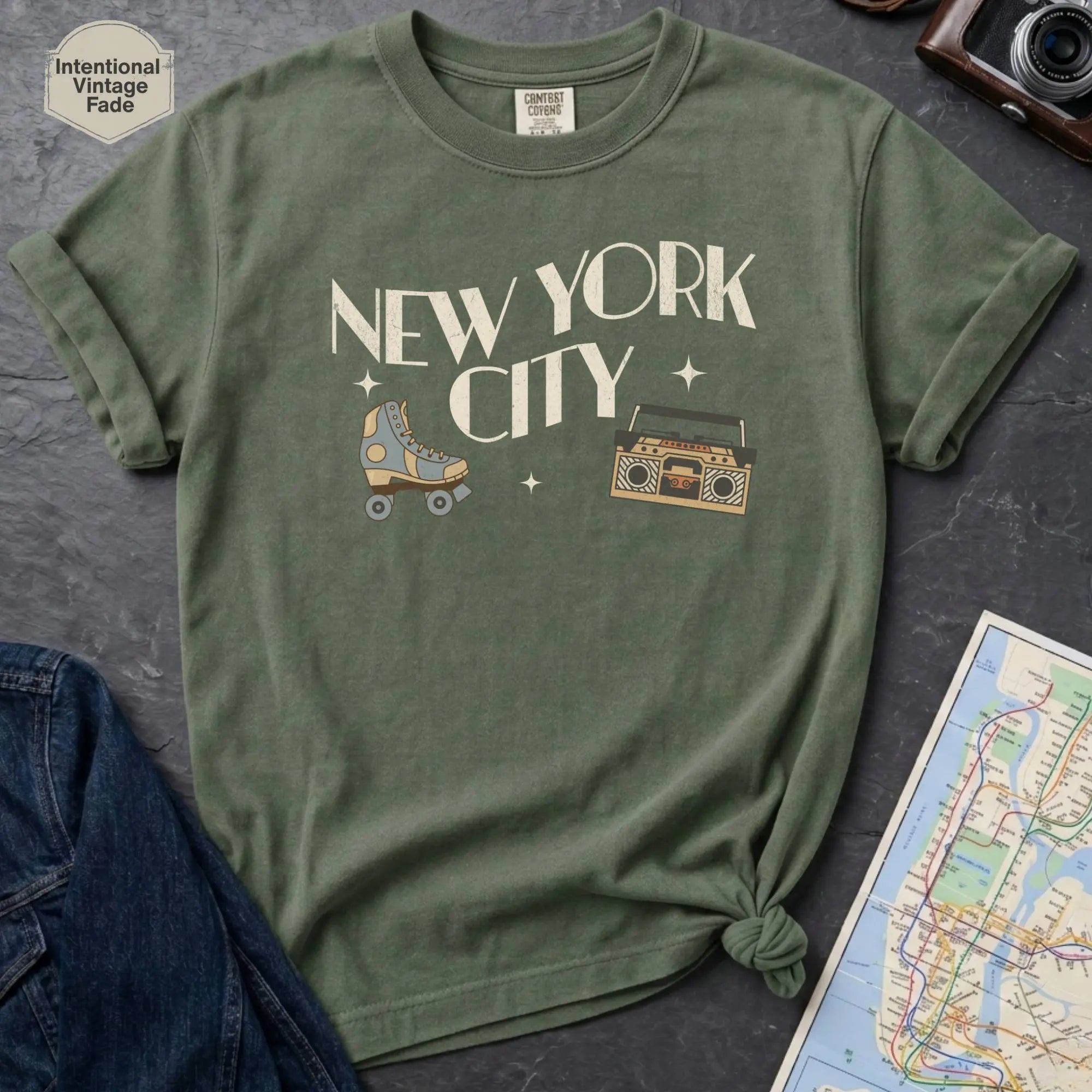 Moss green t-shirt with 'New York City' text and a roller skate and boom box on a dark surface with a map and camera.