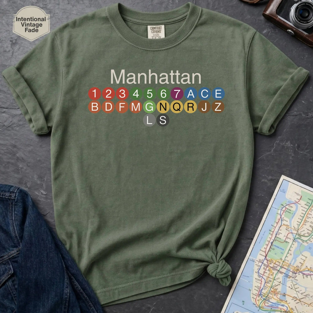 Moss green Comfort Colors t-shirt featuring a minimalist Manhattan subway line graphic with colorful NYC train icons.