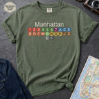 Moss green Comfort Colors t-shirt featuring a minimalist Manhattan subway line graphic with colorful NYC train icons.