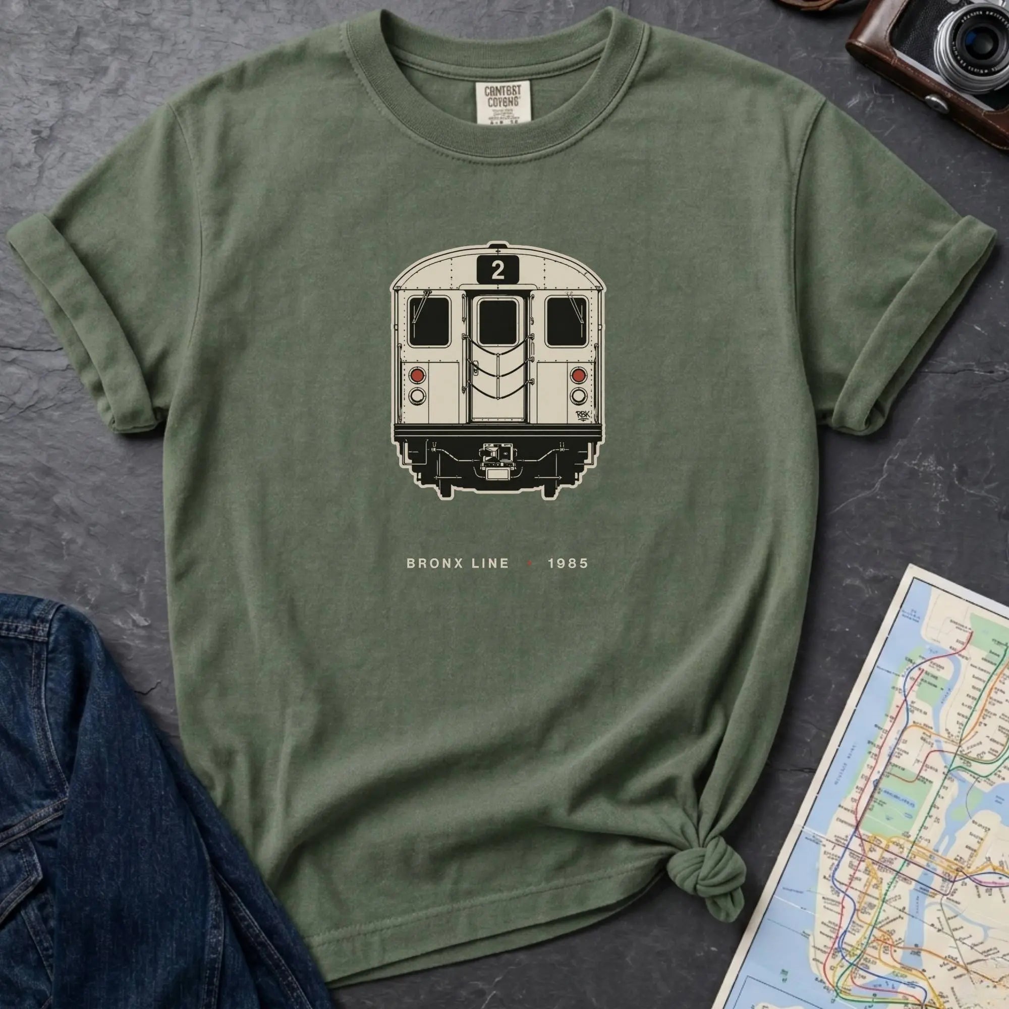 Moss Green t-shirt with a NYC subway train graphic on a dark surface with a map and a denim jacket.