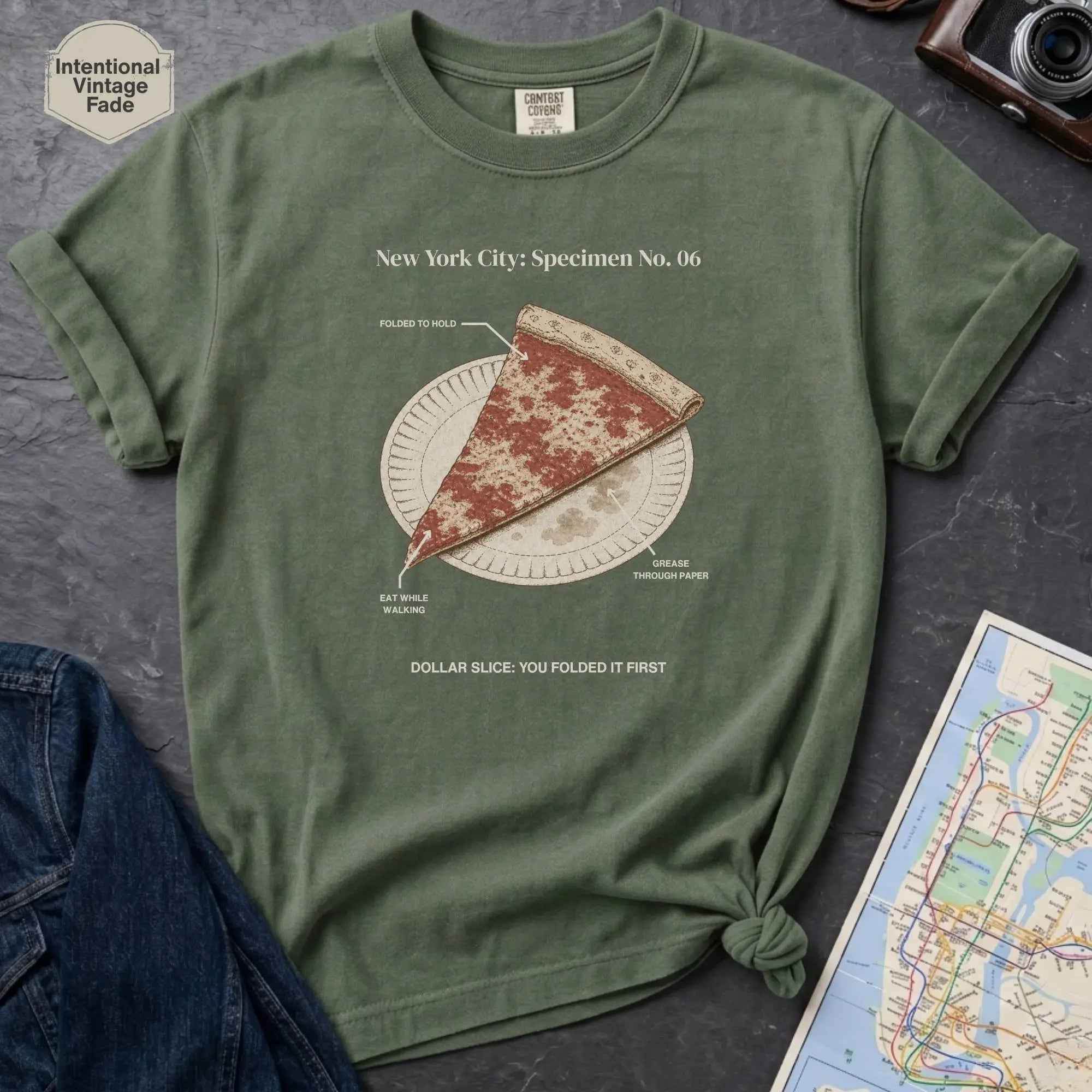 Moss Green Comfort Colors t-shirt featuring a vintage New York City dollar slice pizza graphic with specimen-style callouts and a folded slice on a paper plate.