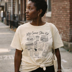 Woman wearing a Ivory Comfort Colors t-shirt with 'NY Corner Store Kid' text and images on a gray surface with denim jacket and map on a NYC street.
