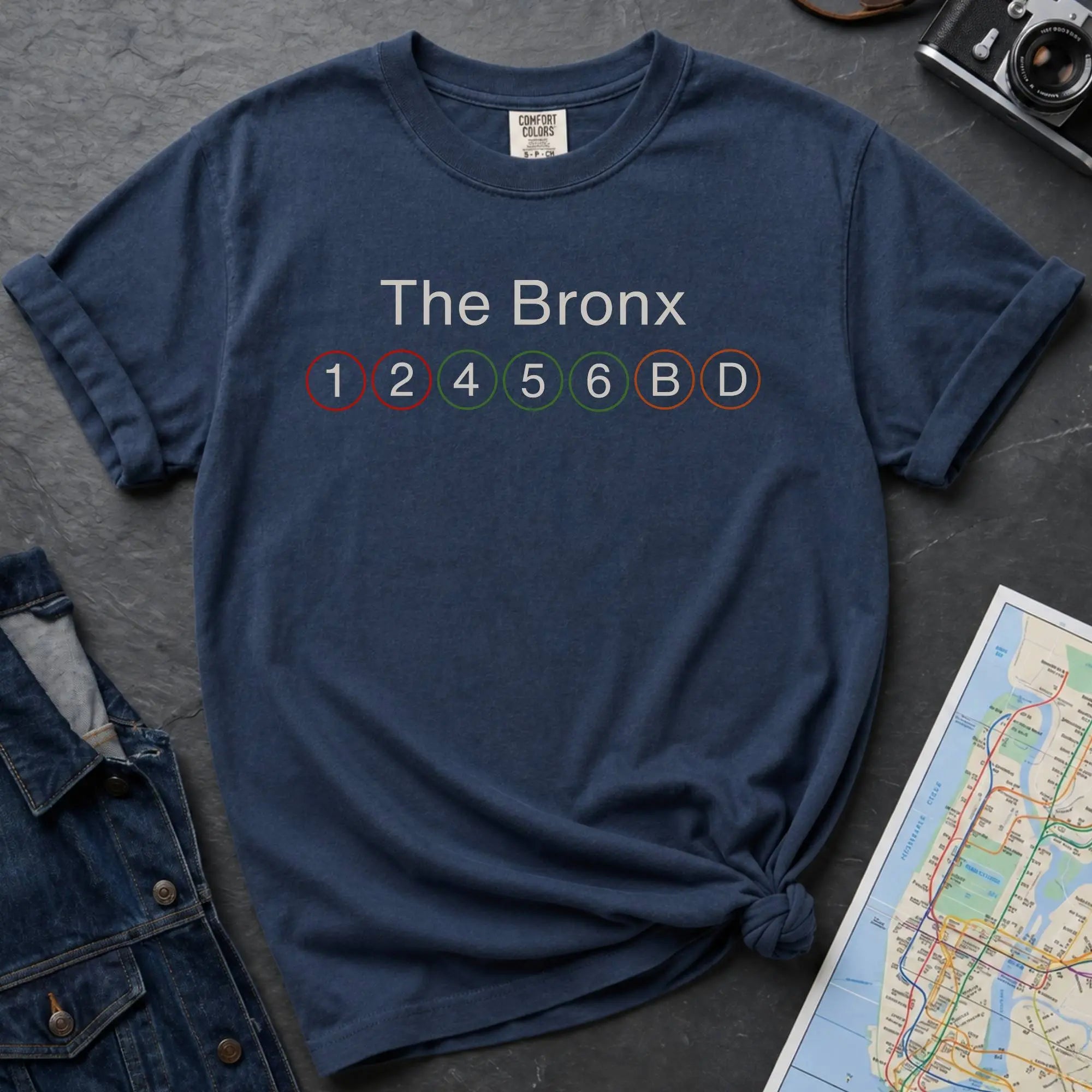 Navy t-shirt with 'The Bronx' and subway line numbers on a dark surface with a map and camera.