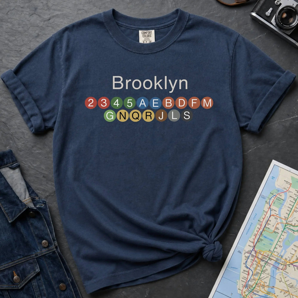 Navy t-shirt with 'Brooklyn' and subway line numbers on a dark surface with a map and camera.