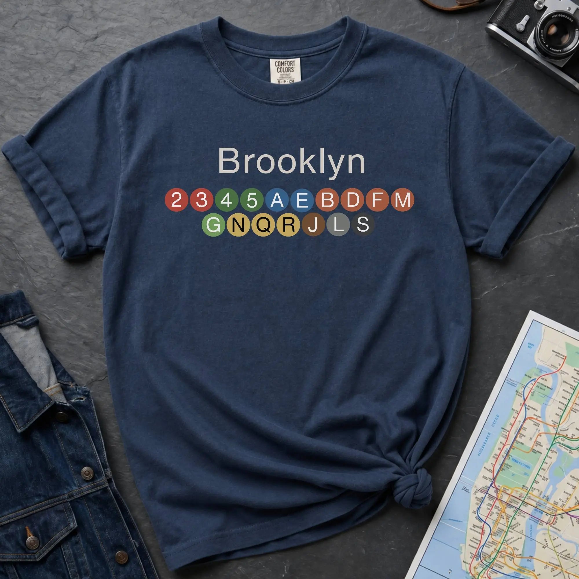 Navy t-shirt with 'Brooklyn' and subway line numbers on a dark surface with a map and camera.