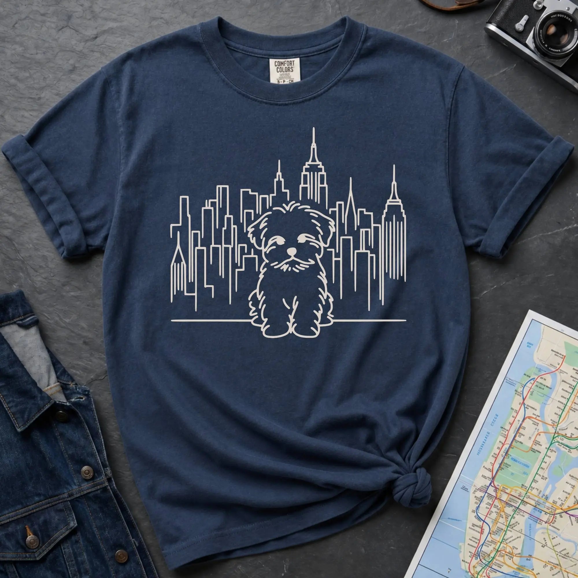 Navy Blue t-shirt with a Maltese dog and cityscape design on a gray surface with a map and camera.