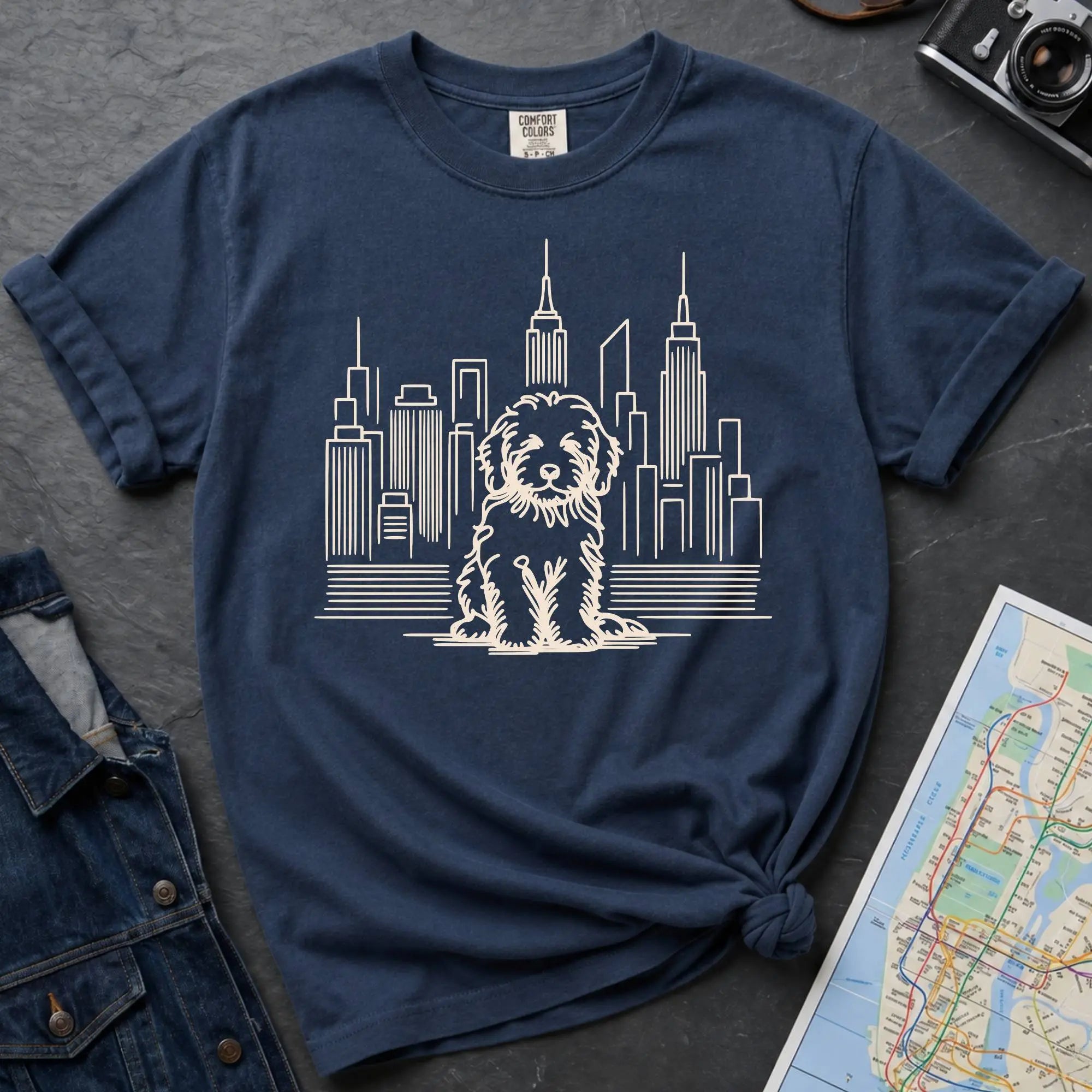 Navy t-shirt with a Golden Doodle and cityscape design on a dark surface with a map and camera.