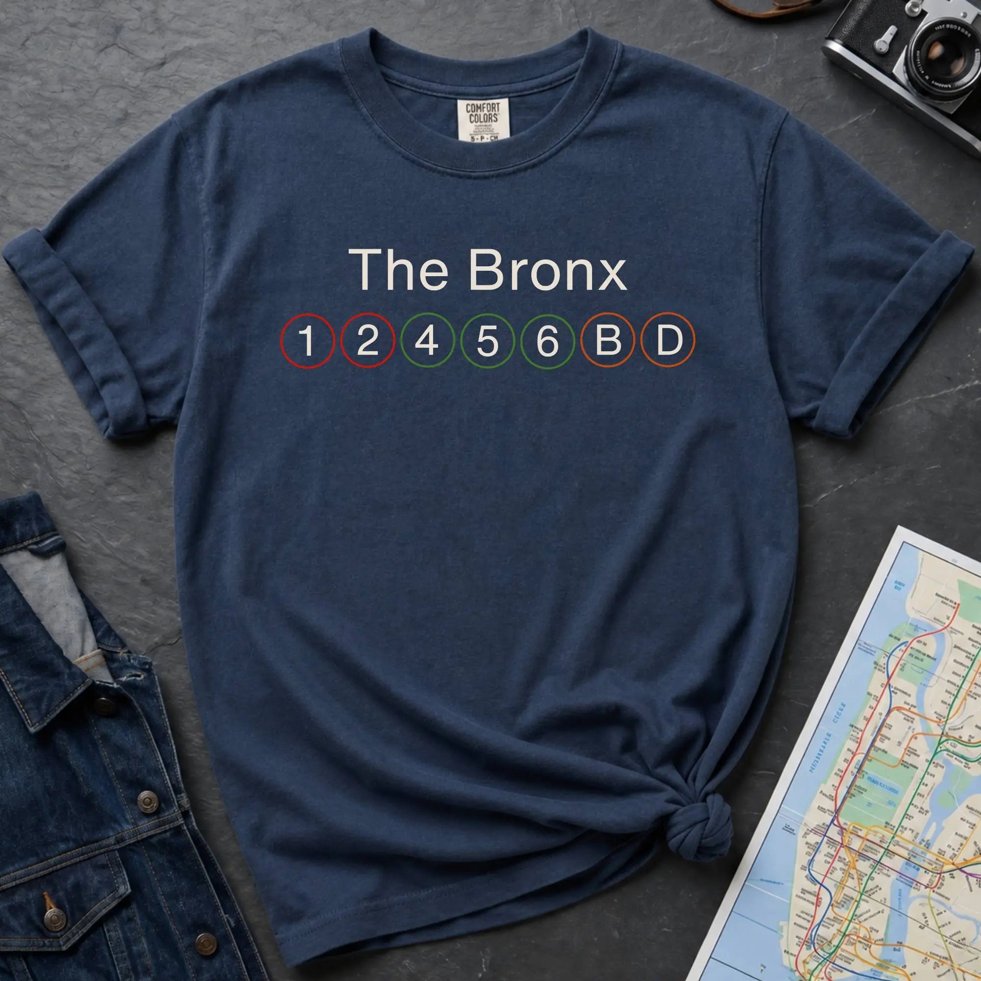 Navy t-shirt with 'The Bronx' and subway line numbers on a dark surface with a map and camera.