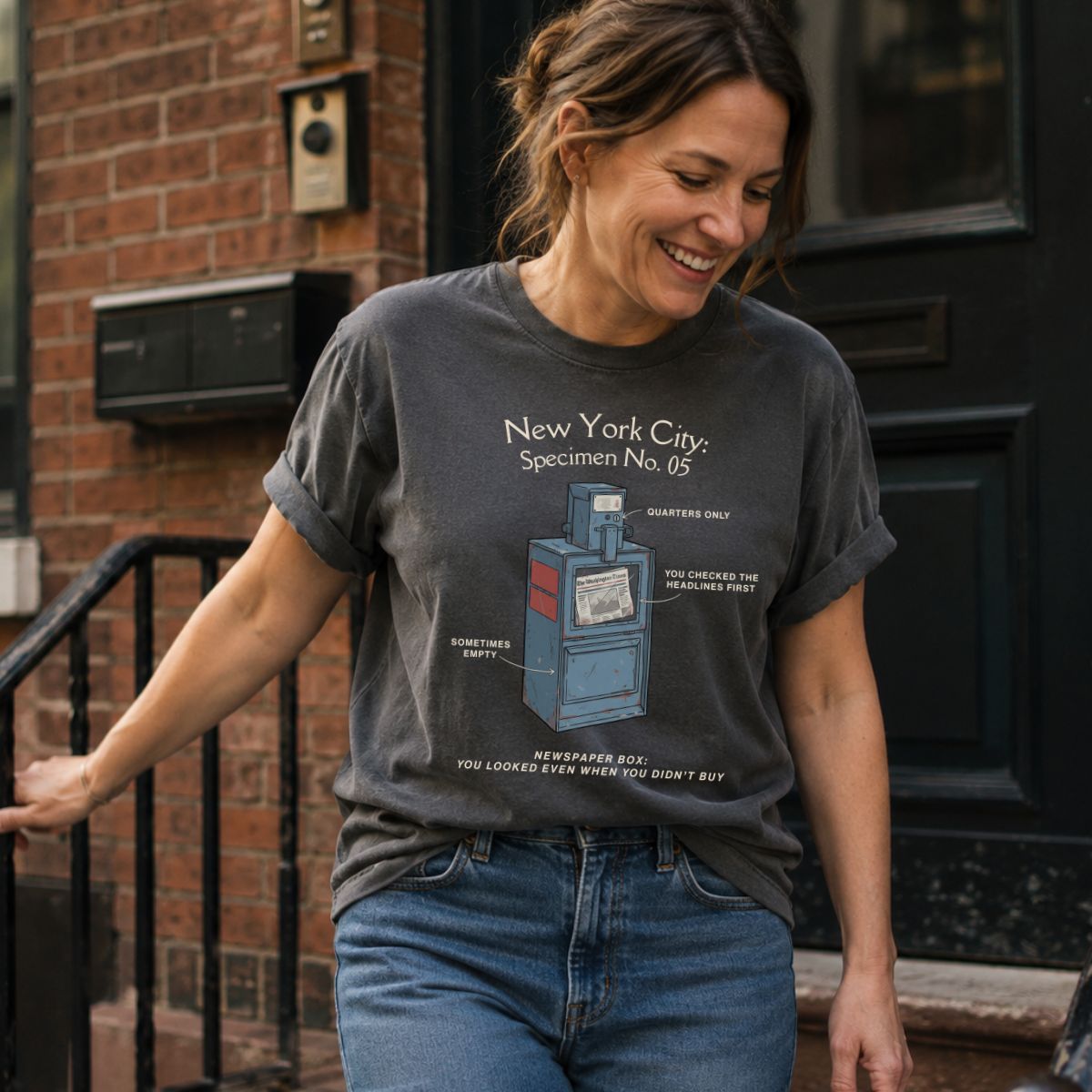 Woman wearing a Pepper colored Comfort Colors t-shirt with a graphic design of a newspaper box in New York City.