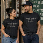 Two people wearing black Comfort Colors t-shirts with coordinates and the text 'our first kiss', standing in front of a store.