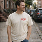 A man wearing an ivory Comfort Colors T-shirt with red Pearl Paint NYC small front chest print, while standing on a NYC street.