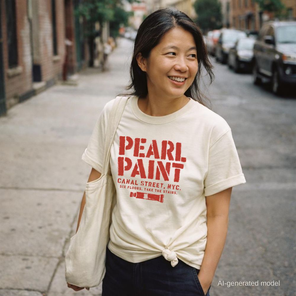 Woman wearing an ivory Comfort Colors t-shirt with a paint tube graphic and 'Pearl Paint' in large distressed stencil lettering, Canal Street NYC and Six Floors Take The Stairs below, standing on a city street with a tote bag.