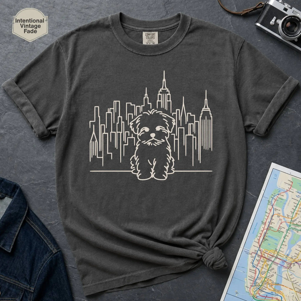 Pepper colored t-shirt with a Maltese dog and cityscape design on a gray surface with a map and camera.