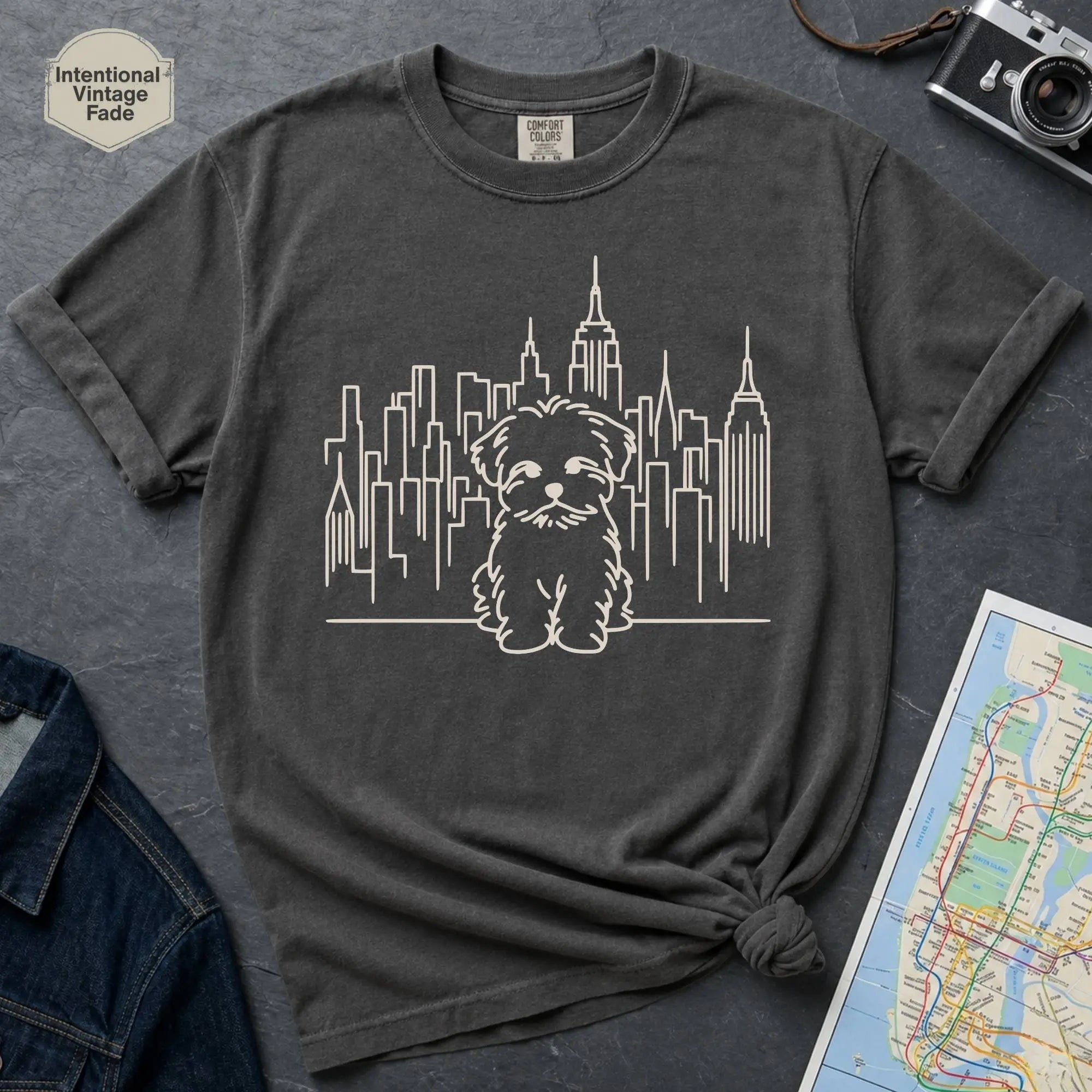 Pepper colored t-shirt with a Maltese dog and cityscape design on a gray surface with a map and camera.
