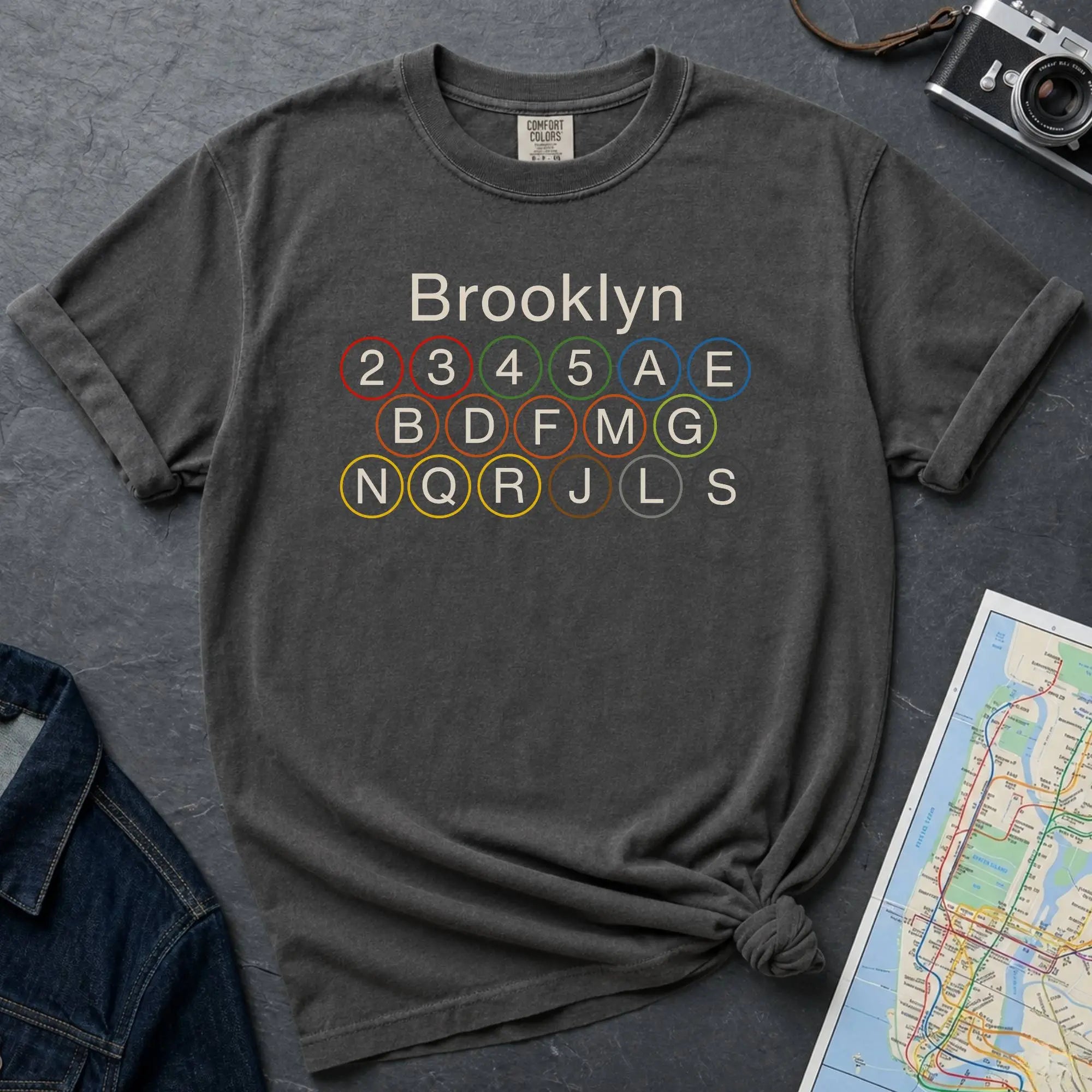 Pepper colored Comfort Colors t-shirt featuring the word “Brooklyn” above colorful NYC subway line circles representing the subway routes that run through Brooklyn.