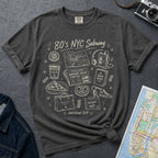 Pepper colored t-shirt with '80s NYC Subway Survival Kit' graphic on a dark surface with a map and jeans.