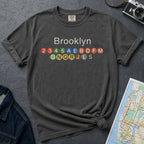 Pepper colored t-shirt with 'Brooklyn' and subway line numbers on a dark surface with a map and camera.