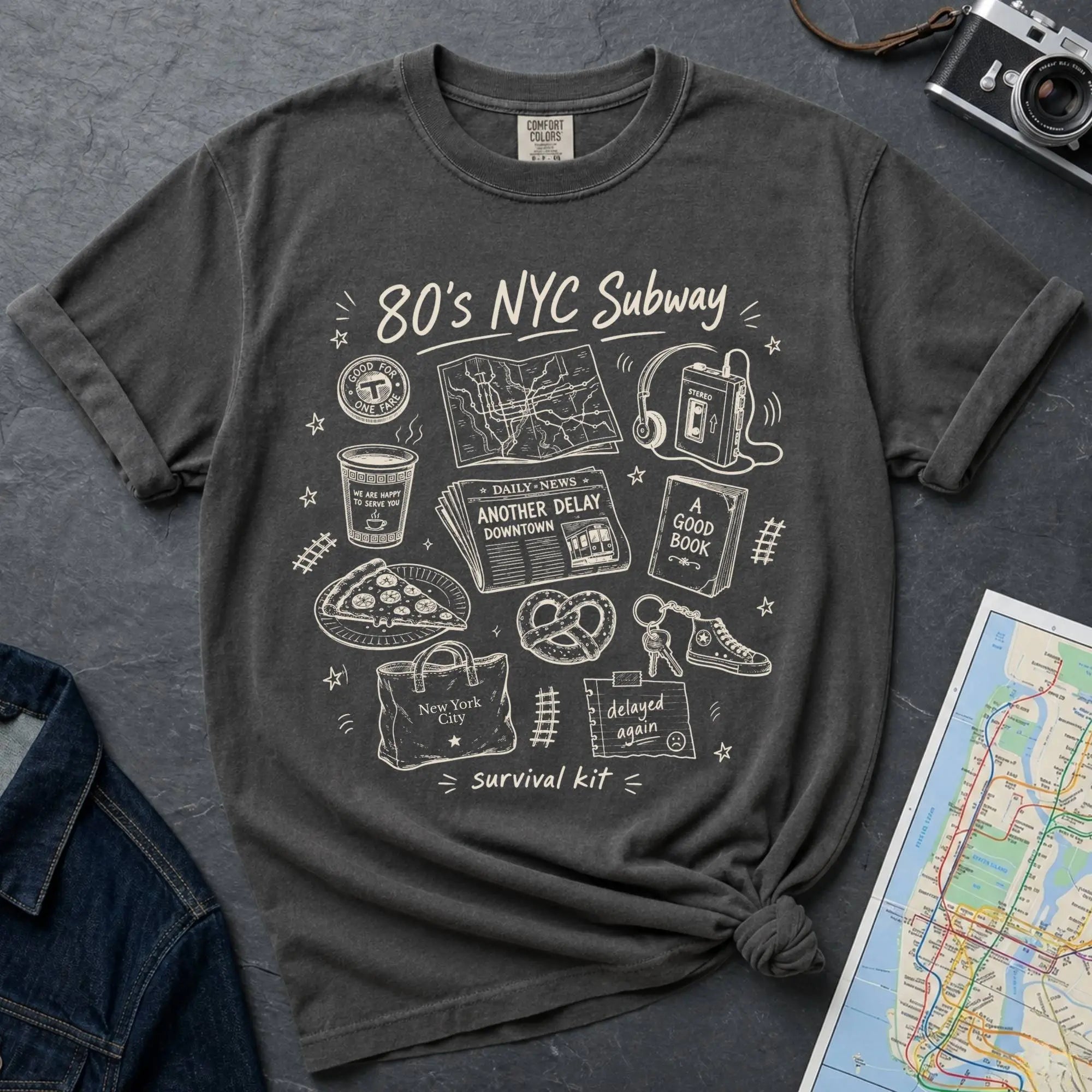 Pepper colored t-shirt with '80s NYC Subway Survival Kit' graphic design on a dark surface with a map and denim jacket.