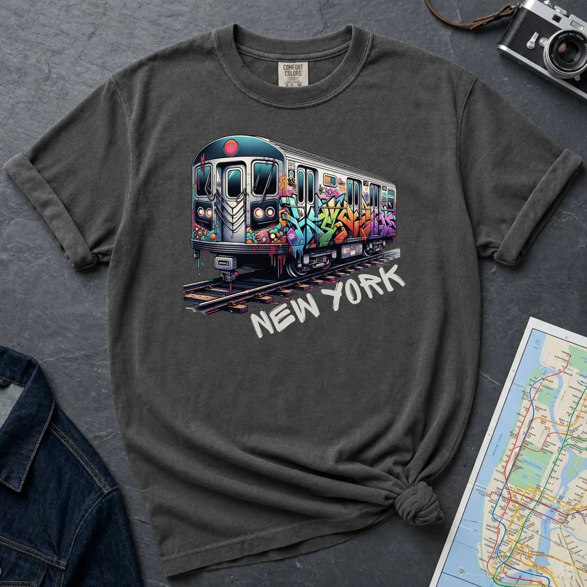 Pepper colored t-shirt with a colorful train graphic and 'New York' text on a gray surface with a map and denim jacket.