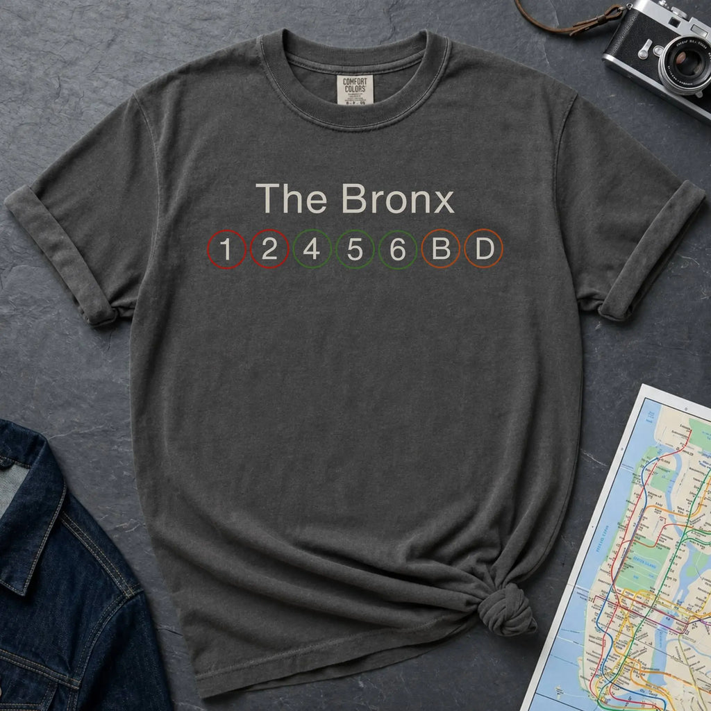 Pepper colored t-shirt with 'The Bronx' and subway line numbers on a dark surface with a map and camera.