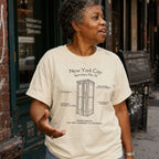 Woman wearing an ivory colored Comfort Colors t-shirt with text and a vintage phone booth design, standing on a street.