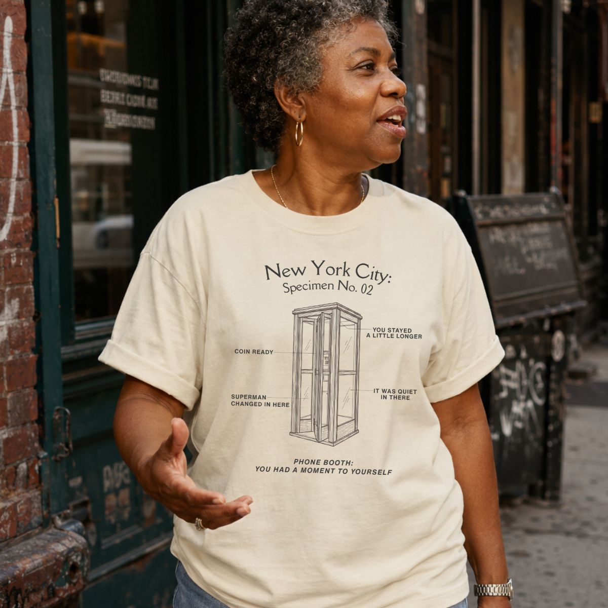 Woman wearing an ivory colored Comfort Colors t-shirt with text and a vintage phone booth design, standing on a street.
