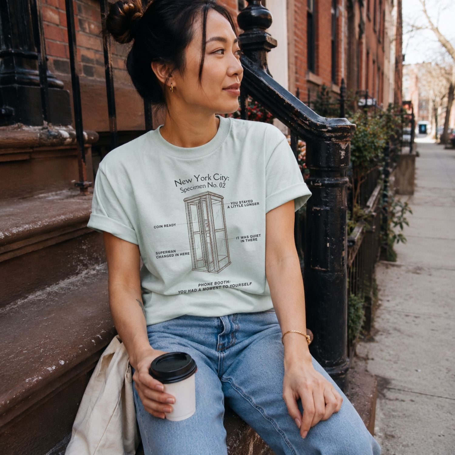 A woman wearing a Bay colored Comfort Colors t-shirt featuring a vintage New York City phone booth graphic with specimen-style callouts and “Phone Booth: You Had a Moment to Yourself” text.