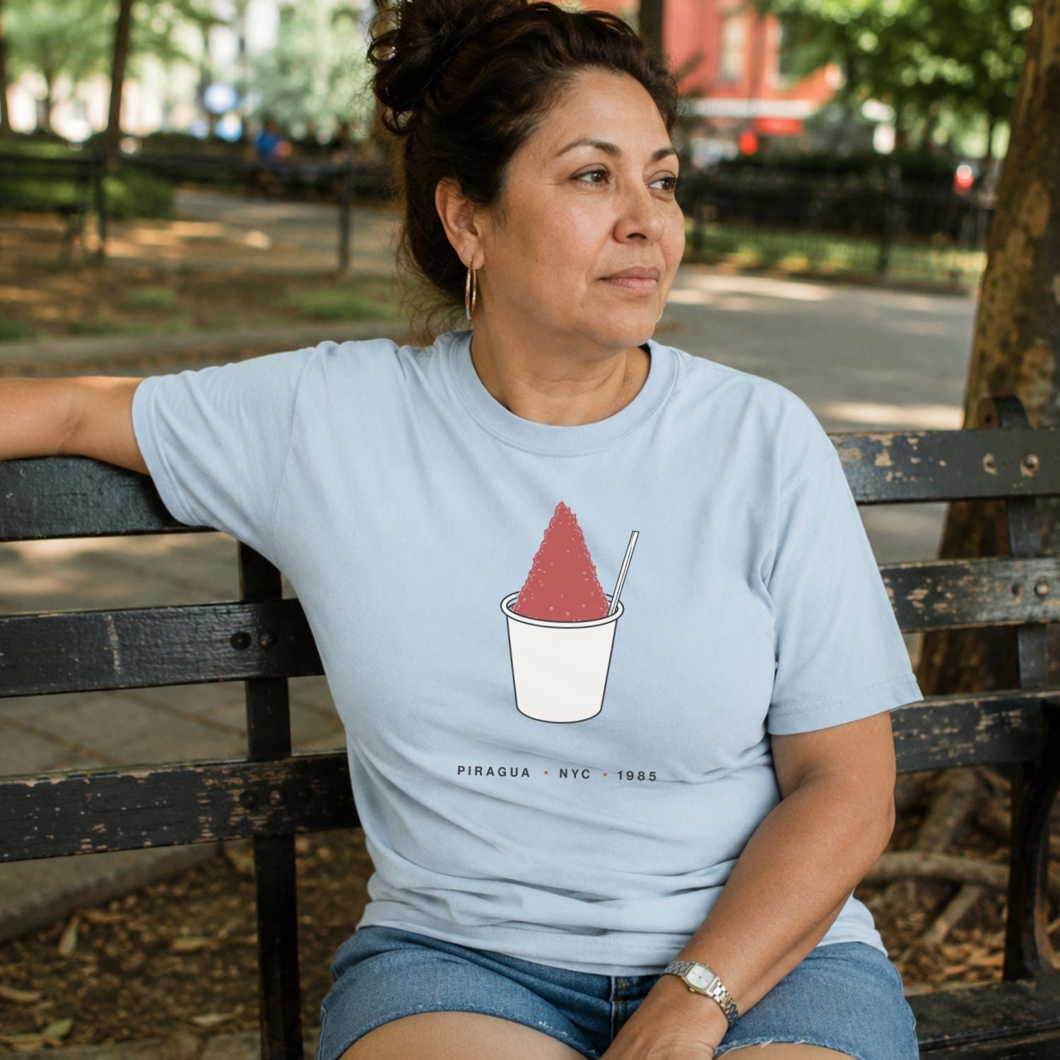 Woman sitting on a bench wearing a light blue Comfort Colors t-shirt with a piragua graphic and text, placed on a dark surface with a map and denim jacket.