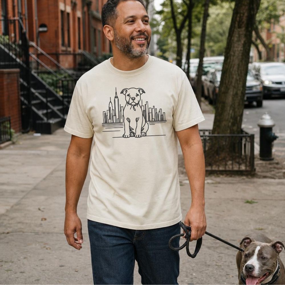 Man walking a dog on a leash in an urban setting, wearing an Ivory Comfort Colors t-shirt with a Pitbull dog and cityscape design.