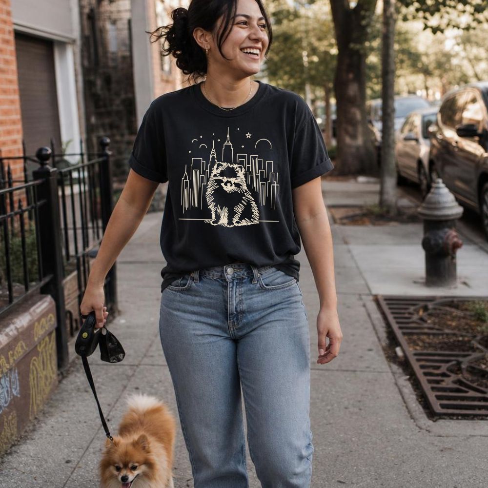 Woman walking a Pomeranian on a sidewalk wearing a black Comfort Colors t-shirt featuring a fluffy Pomeranian dog illustration in front of the New York City skyline.