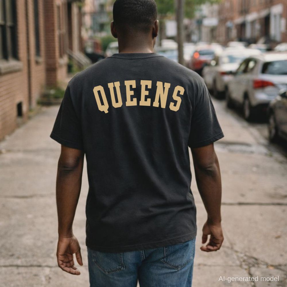 Man wearing a black Comfort Colors t-shirt with 'Queens' printed on the back, standing on a street.