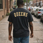 Man wearing a black Comfort Colors t-shirt with 'Queens' printed on the back, standing on a street.