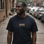 Man wearing a black Comfort Colors t-shirt with 'Queens New York City' text on a street.