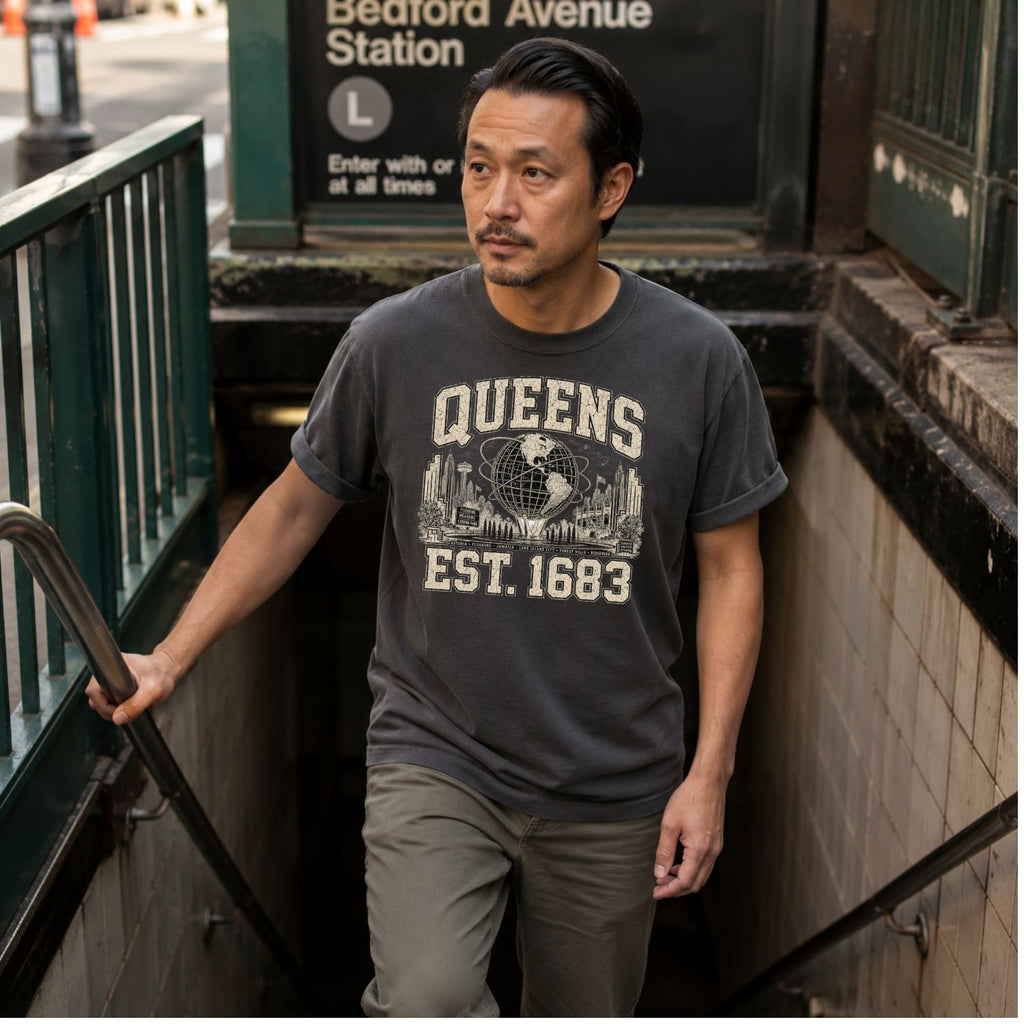 Man wearing a 'Queens Est. 1683' t-shirt standing on a subway platform.