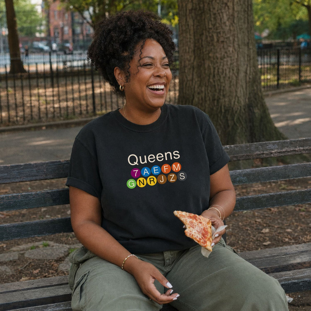 Woman sitting on a bench in a park wearing a black Comfort Colors t-shirt featuring a minimalist Queens subway line graphic with colorful NYC train icons while holding a slice of pizza.