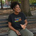 Woman sitting on a bench in a park wearing a black Comfort Colors t-shirt featuring a minimalist Queens subway line graphic with colorful NYC train icons while holding a slice of pizza.