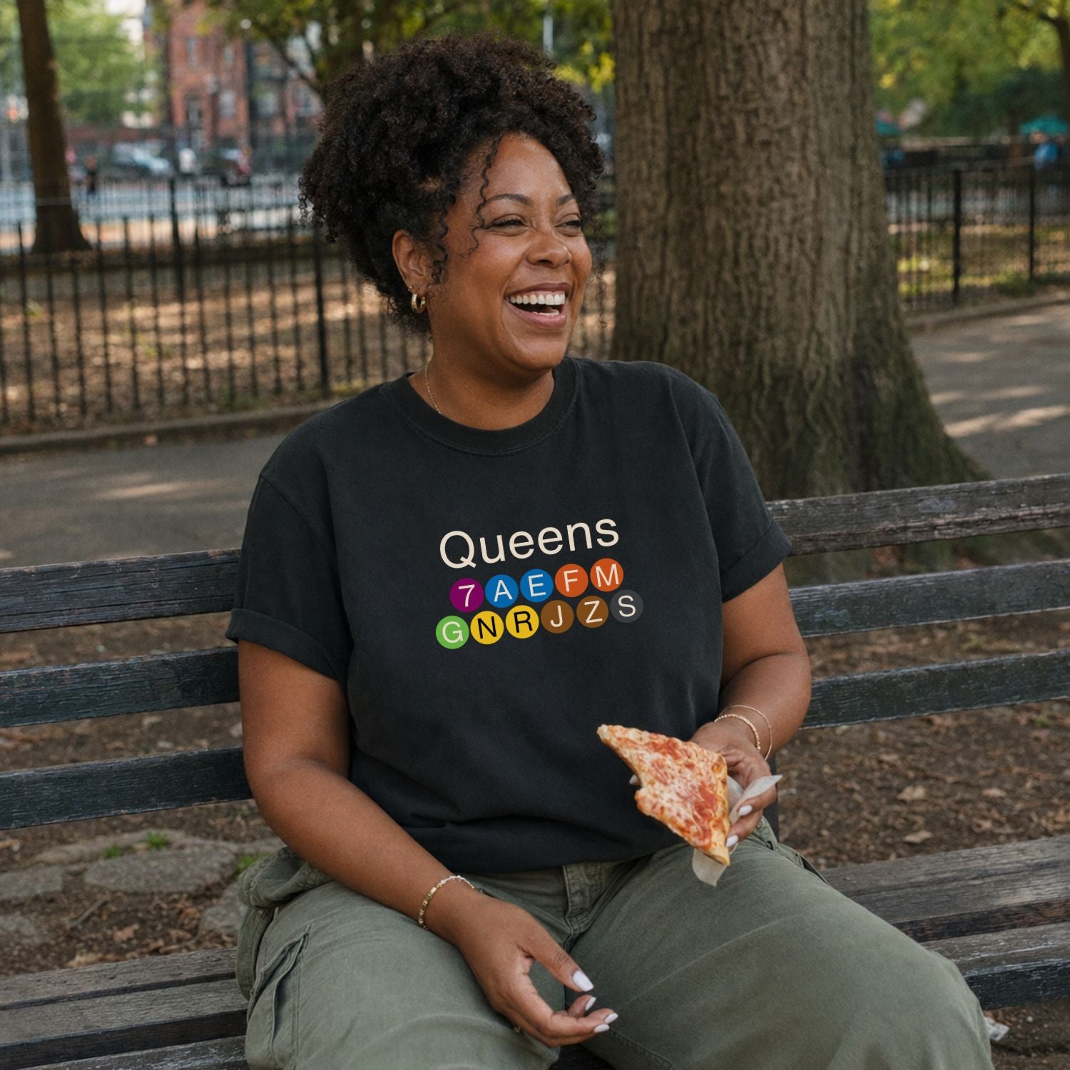 Woman sitting on a bench in a park wearing a black Comfort Colors t-shirt featuring a minimalist Queens subway line graphic with colorful NYC train icons while holding a slice of pizza.