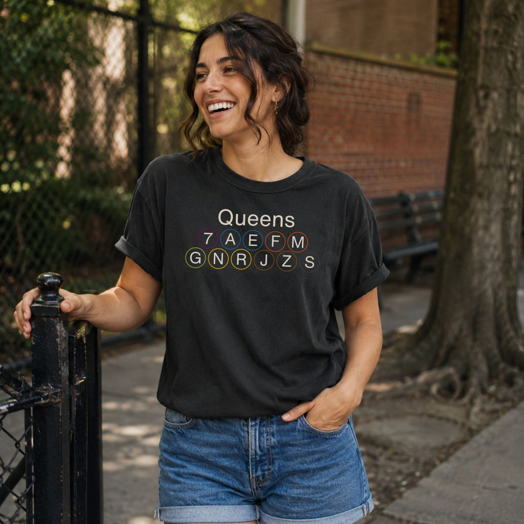 Woman wearing a Black Comfort Colors t-shirt featuring a minimalist Queens subway line graphic with Queens text and classic NYC train icons standing outdoors near a tree and bench.