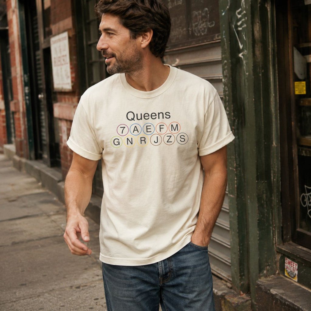 Man wearing a Ivory t-shirt with 'Queens' and subway line numbers standing on a street.