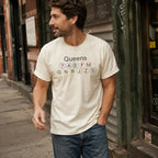 Man wearing a Ivory t-shirt with 'Queens' and subway line numbers standing on a street.