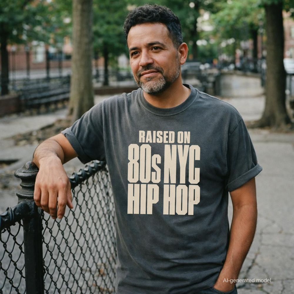 Man wearing a pepper colored Comfort Colors T-shirt with cream Raised on 80s NYC Hip Hop front print, leaning on a fence in a city park. 
